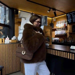 Op de afbeelding: Een vrouw draagt een bruin nep bontjasje, grijze sweatpants en een bruine su&egrave;de handtas met een gouden hoefijzer gesp. Ze staat in een koffiebar met een zwarte toonbank en houten lattenwanden. De tekst "DAILY ROSE COFFEE" is zichtbaar op de toonbank.