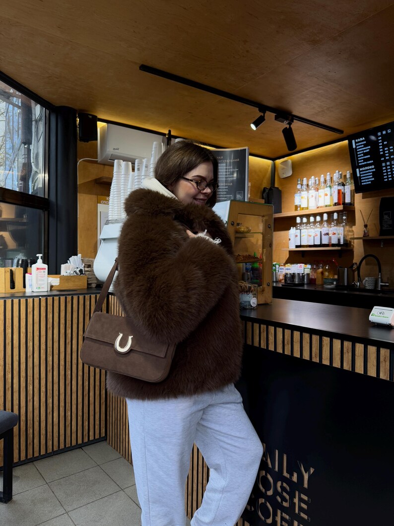 Op de afbeelding: Een vrouw draagt een bruin nep bontjasje, grijze sweatpants en een bruine su&egrave;de handtas met een gouden hoefijzer gesp. Ze staat in een koffiebar met een zwarte toonbank en houten lattenwanden. De tekst "DAILY ROSE COFFEE" is zichtbaar op de toonbank.