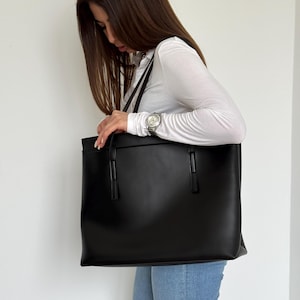 May include: A large, black tote bag with two handles and a rectangular shape. The bag is made of a smooth material and has a minimalist design. The bag is being held by a person wearing a white shirt and blue jeans.
