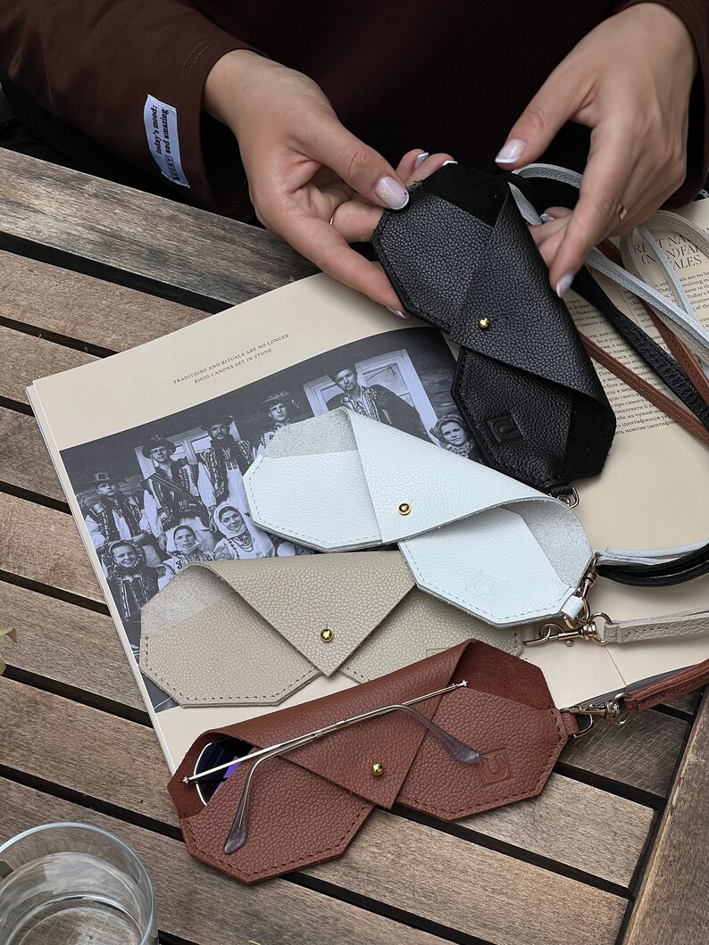 May include: Four different leather eyeglass cases in various colors, including brown, beige, white, and black. The cases are folded in a triangular shape and have a gold metal clasp. The cases are arranged on a wooden surface.