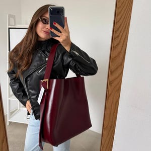 Peut inclure: Un sac à épaule bordeaux profond avec une finition lisse et structurée, doté d'une boucle en métal doré sur la large bandoulière. Le sac présente un design épuré avec une étiquette rectangulaire blanche attachée sur le côté près de la boucle.