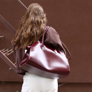 Op de afbeelding: Een grote, bordeauxrode leren tote bag met een gestructureerd ontwerp. De tas heeft een glanzende afwerking en een handvat aan de bovenkant en wordt over de schouder gedragen. De persoon draagt een bruine jas en een witte broek.
