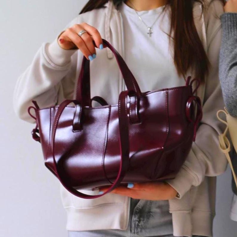 May include: A burgundy leather handbag with a glossy finish. The bag has two short handles and a longer shoulder strap. The design includes a structured shape with visible stitching and a circular detail on the sides.