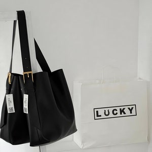 May include: A black leather tote bag with gold buckles and two white tags that say "LUCKY" hanging from the handles. The bag is sitting on a white surface next to a white paper bag with the word "LUCKY" printed in black.