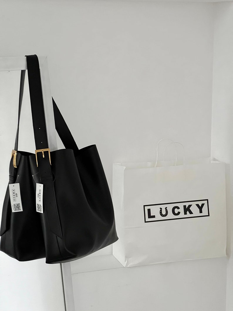 May include: A black leather tote bag with gold buckles and two white tags that say "LUCKY" hanging from the handles. The bag is sitting on a white surface next to a white paper bag with the word "LUCKY" printed in black.