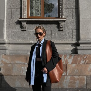 May include: A person wearing a black blazer over a light blue button-down shirt and black pants. They are carrying a brown leather tote bag and wearing sunglasses.