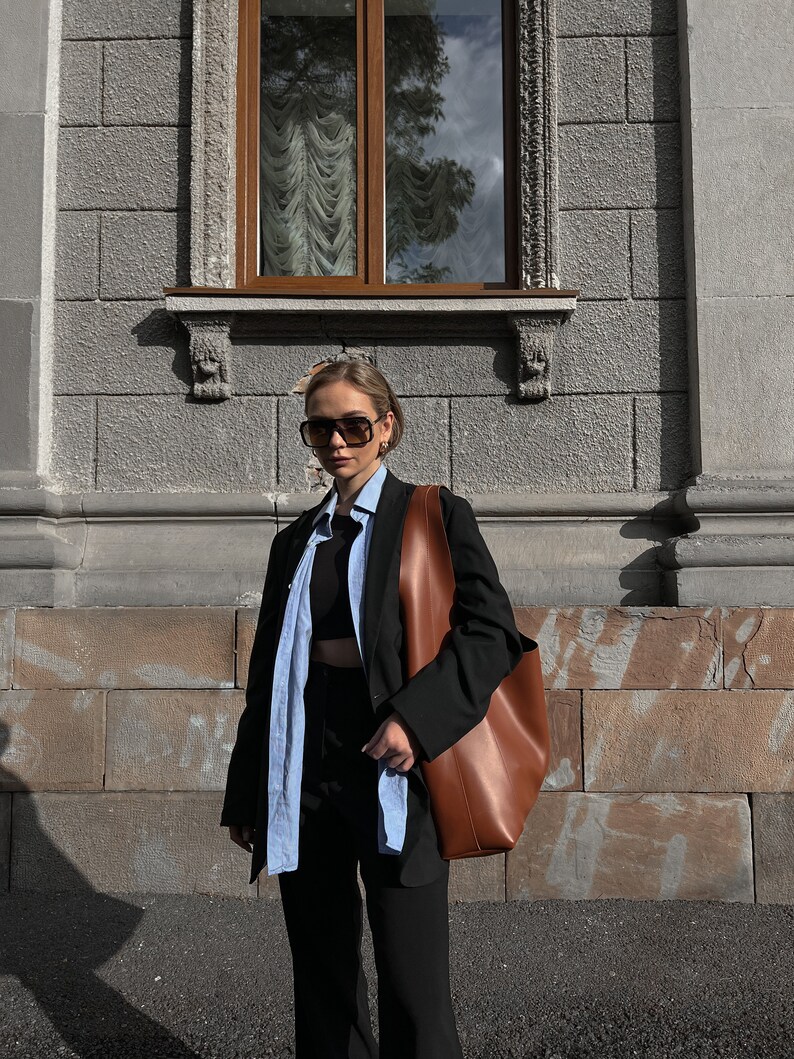 May include: A person wearing a black blazer over a light blue button-down shirt and black pants. They are carrying a brown leather tote bag and wearing sunglasses.