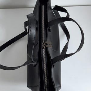 May include: Black tote bag with a zipper closure and two handles. The bag is made of a smooth material and has a rectangular shape. The zipper is silver-colored, and the bag appears to be empty.