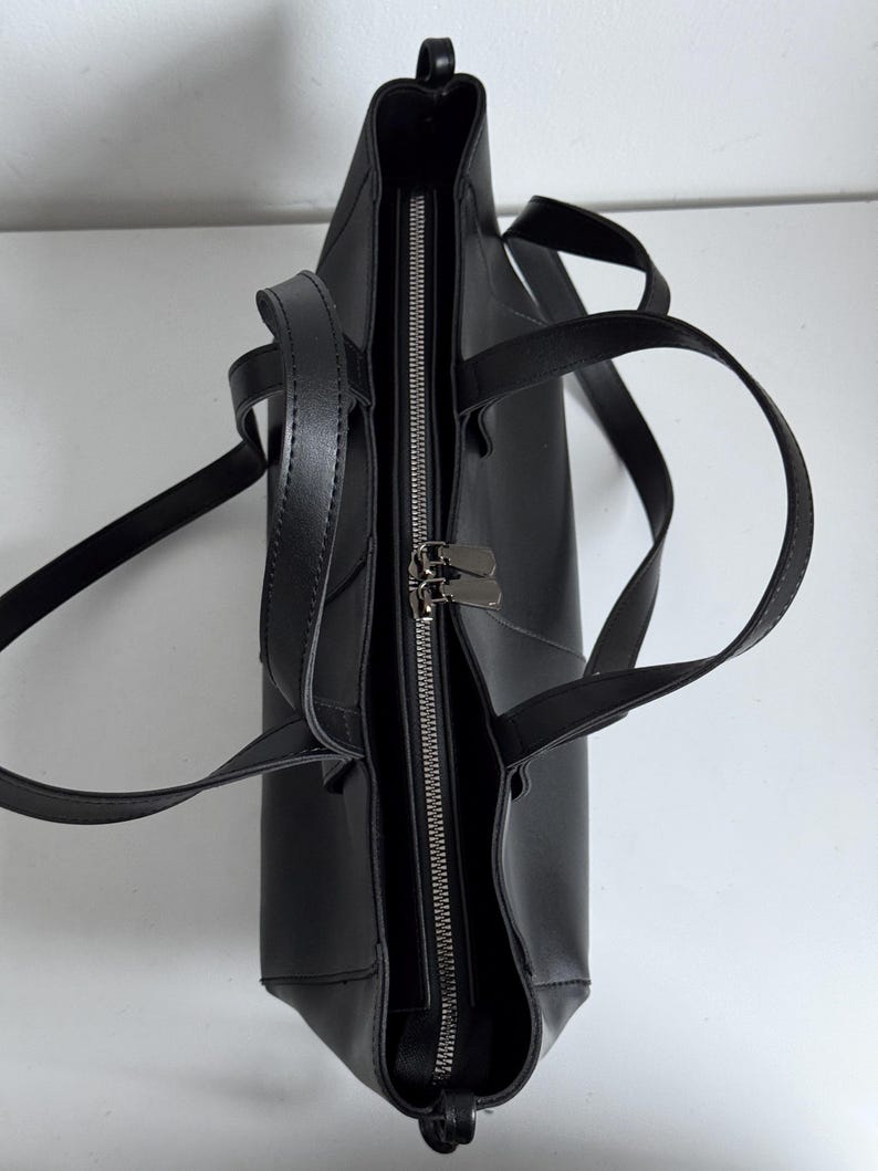 May include: Black tote bag with a zipper closure and two handles. The bag is made of a smooth material and has a rectangular shape. The zipper is silver-colored, and the bag appears to be empty.