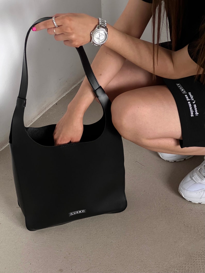 May include: A black shoulder bag with a rectangular shape and a long strap. The bag has a minimalist design with the word "LUCKY" in a small, rectangular silver label. The bag is held by a person wearing a watch and rings.