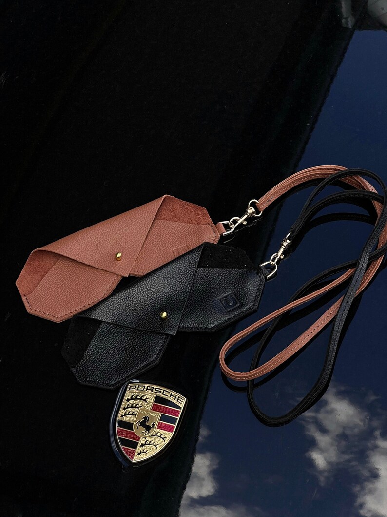 May include: Two leather eyeglass cases, one brown and one black, with a brown and black lanyard. The cases are folded and have a gold metal clasp. The lanyard has a gold metal clip. The cases are on a black surface with a Porsche logo reflected in the surface.