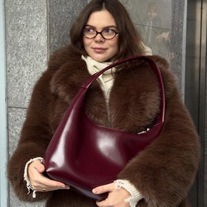 Peut inclure: Un sac à bandoulière de style hobo, d'un bordeaux profond et lisse, présentant une silhouette incurvée et une anse unique intégrée. Le sac arbore un design épuré avec une finition polie et une fermeture éclair sécurisée sur le dessus.