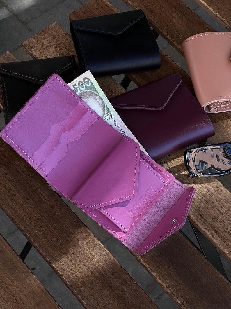 May include: A pink leather wallet with a flap closure and the word "LUCKY" embossed on the front. The wallet is open and there is a bill of Ukrainian currency inside.