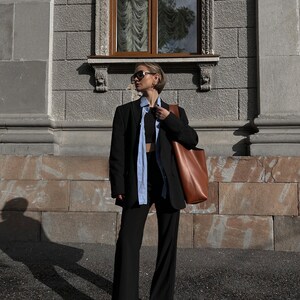 May include: A person wearing a black blazer, a light blue shirt, and black pants. They are carrying a brown leather tote bag.
