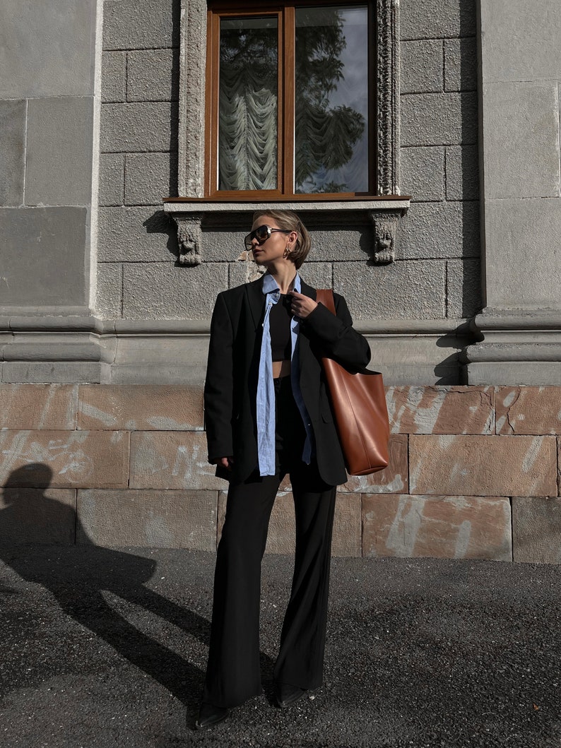 May include: A person wearing a black blazer, a light blue shirt, and black pants. They are carrying a brown leather tote bag.