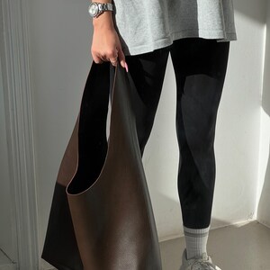 May include: A large brown leather tote bag with a wide opening and a single strap. The bag is being held by a person wearing black leggings and a grey t-shirt.