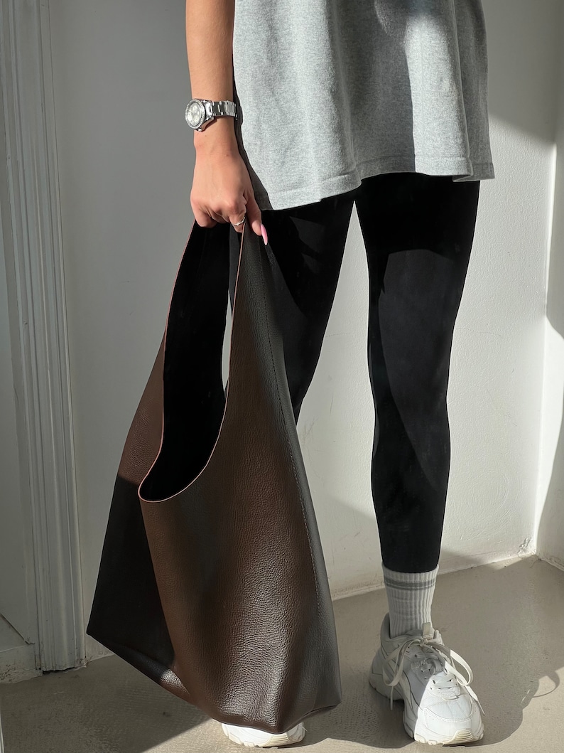 May include: A large brown leather tote bag with a wide opening and a single strap. The bag is being held by a person wearing black leggings and a grey t-shirt.