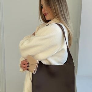 May include: A dark brown leather shoulder bag with a rectangular shape and a long strap. The bag has a small silver "LUCKY" tag on the front. A small beige wallet is also visible.