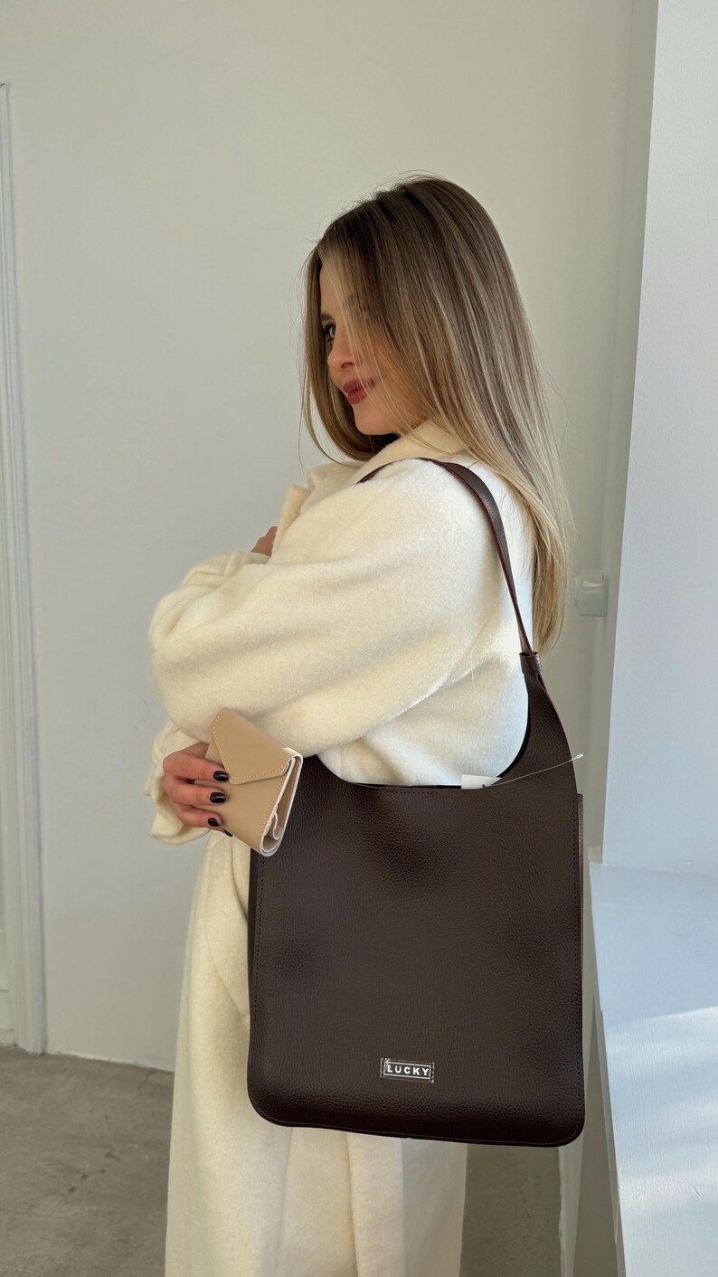 May include: A dark brown leather shoulder bag with a rectangular shape and a long strap. The bag has a small silver "LUCKY" tag on the front. A small beige wallet is also visible.