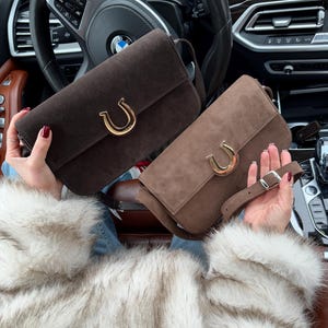 Op de afbeelding: Twee bruine su&egrave;de handtassen met gouden hoefijzervormige sluitingen. De handtassen hebben verstelbare banden en worden getoond in een auto-interieur.