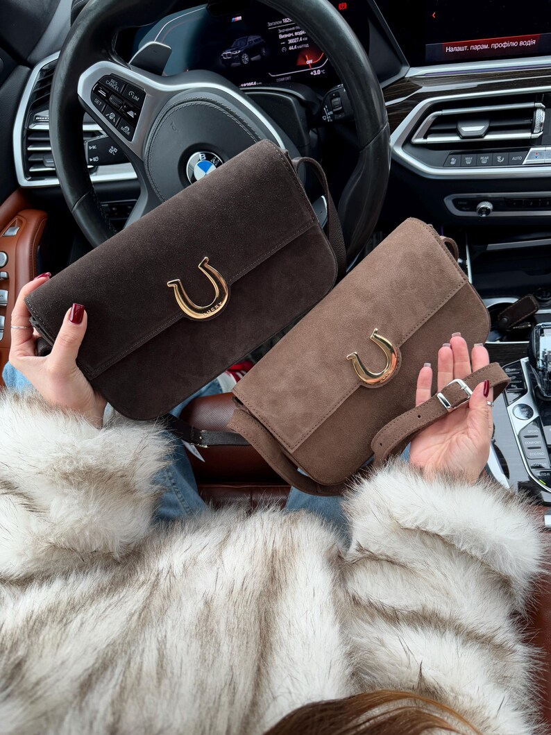Op de afbeelding: Twee bruine su&egrave;de handtassen met gouden hoefijzervormige sluitingen. De handtassen hebben verstelbare banden en worden getoond in een auto-interieur.