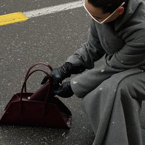 May include: A burgundy handbag with a trapezoid shape and two handles. The bag is on a wet asphalt surface. The person is wearing a gray coat and black gloves.