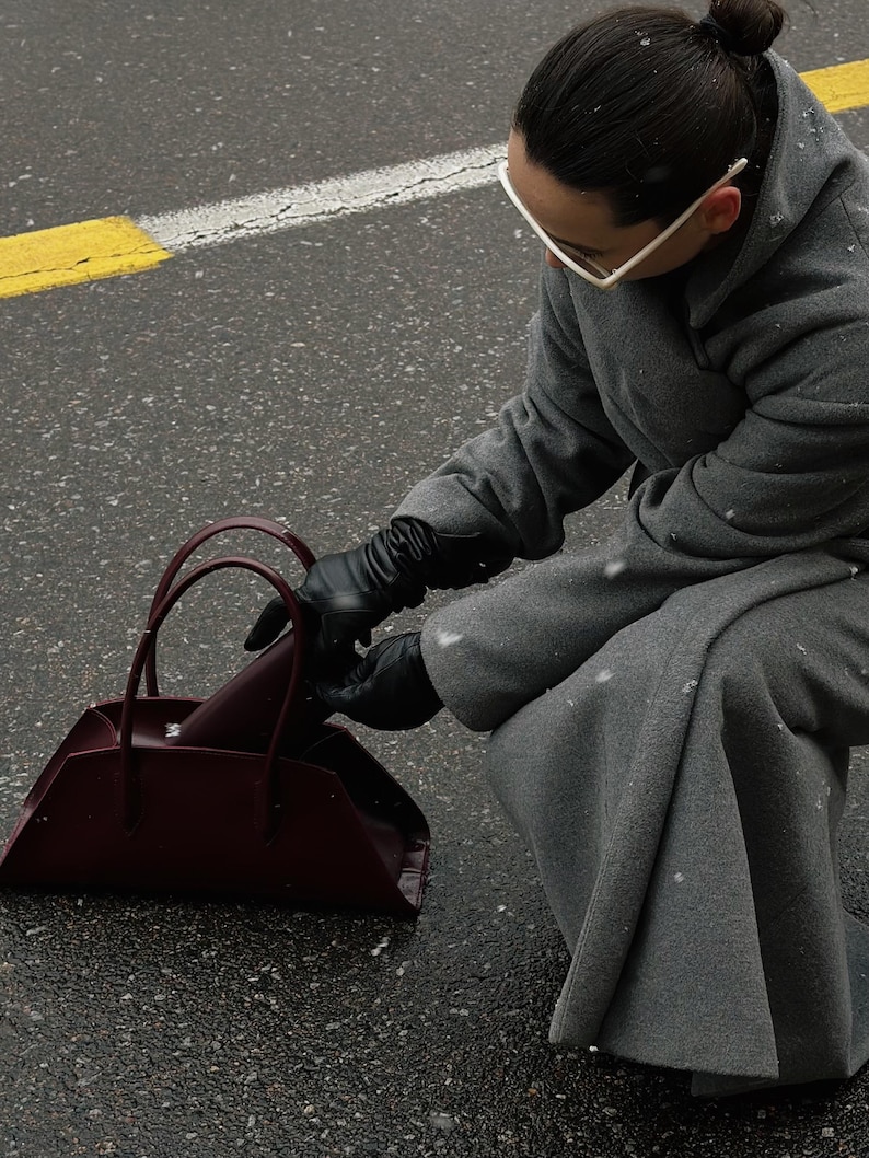 May include: A burgundy handbag with a trapezoid shape and two handles. The bag is on a wet asphalt surface. The person is wearing a gray coat and black gloves.