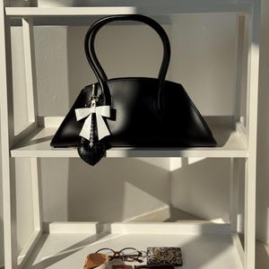 May include: A black handbag with a unique trapezoid shape and curved handles. A white bow charm hangs from the handle. The bag sits on a white shelf with other accessories, including glasses, a small wallet, and a beauty product.