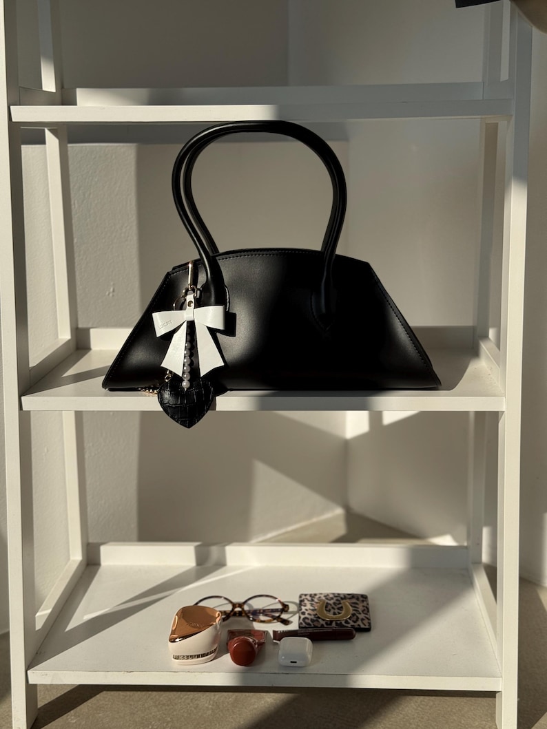 May include: A black handbag with a unique trapezoid shape and curved handles. A white bow charm hangs from the handle. The bag sits on a white shelf with other accessories, including glasses, a small wallet, and a beauty product.