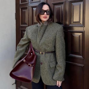 Op de afbeelding: Een groene tweed blazer, bruine riem en bordeauxrode tote bag. De blazer heeft een klassiek ontwerp met een revers en een sluiting met één knoop. De tas heeft een rechthoekige vorm en een gouden gesp.