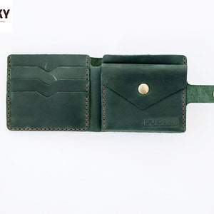 May include: A dark green leather wallet with multiple card slots, a billfold, and a flap pocket. The wallet has a snap closure and a small strap with a button. The wallet is embossed with the word "LUCKY".