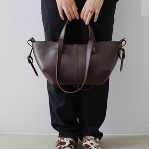 May include: A dark brown leather tote bag with a structured design. The bag has two handles and a shoulder strap. The person is wearing black pants, a gray sweater, and cow-print clogs.