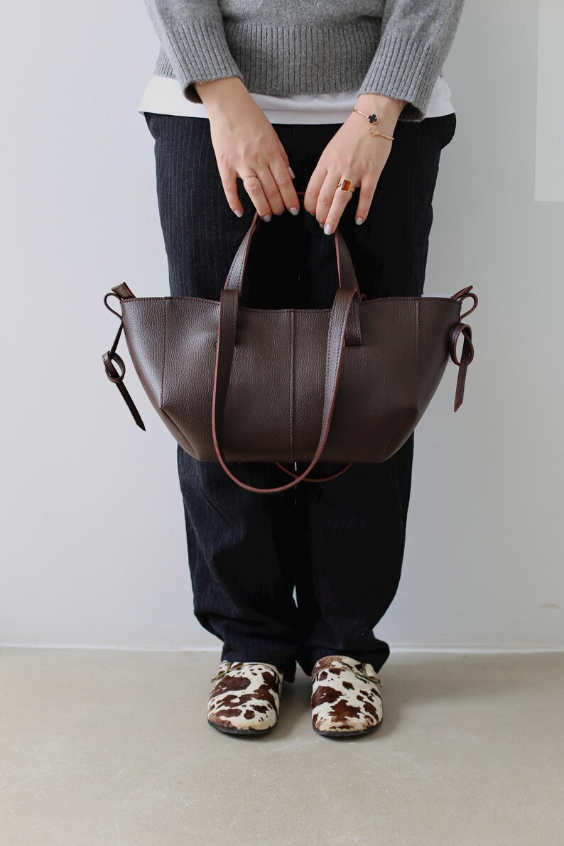 May include: A dark brown leather tote bag with a structured design. The bag has two handles and a shoulder strap. The person is wearing black pants, a gray sweater, and cow-print clogs.