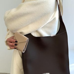May include: A dark brown leather tote bag with a silver "LUCKY" emblem. A small beige wallet is held in the hand. The bag has a long strap and a rectangular shape. The person is wearing a cream-colored coat.
