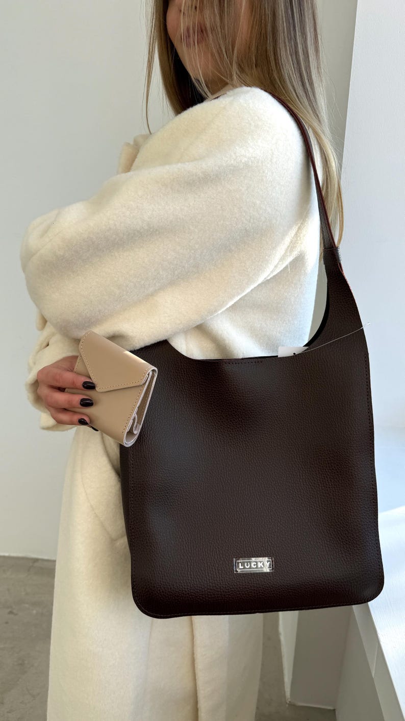 May include: A dark brown leather tote bag with a silver "LUCKY" emblem. A small beige wallet is held in the hand. The bag has a long strap and a rectangular shape. The person is wearing a cream-colored coat.