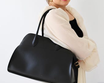 May include: A structured black handbag with a smooth finish, featuring two thin top handles and a rectangular base. The bag has a clean, minimalist design with visible stitching along the top panel and a gold-toned metal buckle on the side strap.