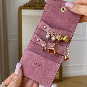 Può includere: Un organizer per gioielli in camoscio rosa polvere con chiusura a bottone a pressione e la parola LUCKY impressa sulla tasca anteriore. L'interno contiene fessure con due orecchini di perle, due orecchini a nodo dorati e due anelli.
