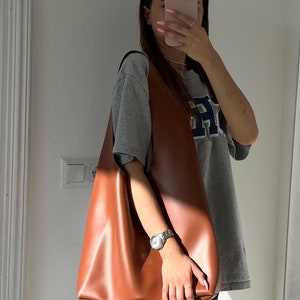 May include: A brown leather tote bag with a slouchy design. The bag is being held by a person wearing a grey t-shirt with a blue and white logo.