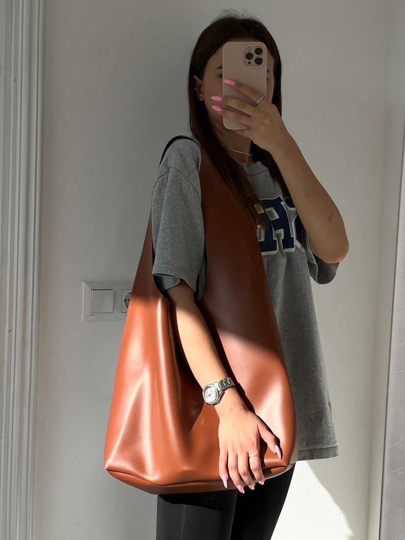 May include: A brown leather tote bag with a slouchy design. The bag is being held by a person wearing a grey t-shirt with a blue and white logo.