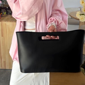 May include: A large, rectangular black tote bag with a smooth, matte finish and a wide, integrated cutout handle at the top center. The bag features a structured, minimalist design with clean lines and a solid black color throughout the exterior.