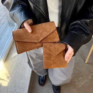 May include: A brown suede envelope-style wallet and a matching smaller square suede coin purse. Both items feature a triangular flap closure and a soft, textured finish. The larger wallet has a subtle embossed logo on the front right corner.