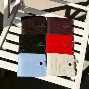 Puede incluir: Seis carteras rectangulares dispuestas en dos columnas. La columna izquierda presenta una cartera marrón oscuro con textura de cocodrilo, una verde oscuro texturizada y una azul claro lisa. La columna derecha muestra una morada profunda, una roja brillante y una crema con textura de cocodrilo. Cada cartera incluye una pequeña lengüeta de cierre asegurada con un botón de presión metálico.