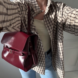 May include: A deep burgundy handbag with a smooth, glossy finish, featuring a structured flap closure and a horizontal strap secured by a silver-toned metal buckle. The bag includes a single vertical handle attached to the top, providing a compact and polished appearance for carrying essentials.