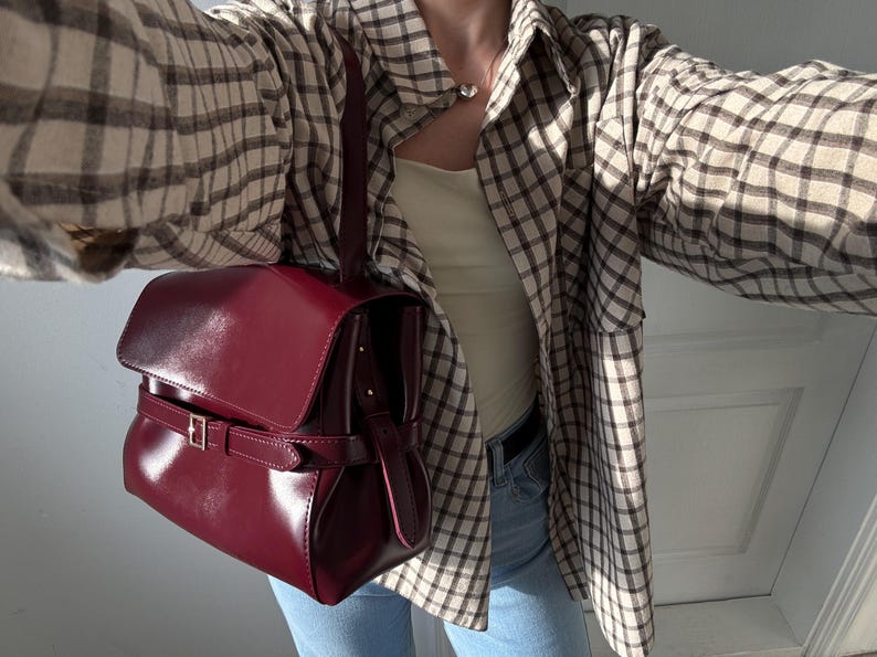 May include: A deep burgundy handbag with a smooth, glossy finish, featuring a structured flap closure and a horizontal strap secured by a silver-toned metal buckle. The bag includes a single vertical handle attached to the top, providing a compact and polished appearance for carrying essentials.