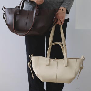 May include: Two leather tote bags in the image. One is a dark brown color with a textured surface and long handles. The other is a cream color with a smooth surface and a zipper closure. Both bags have a simple, elegant design.