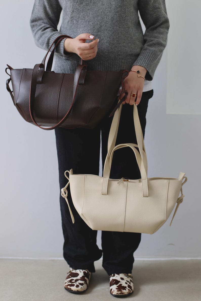 May include: Two leather tote bags in the image. One is a dark brown color with a textured surface and long handles. The other is a cream color with a smooth surface and a zipper closure. Both bags have a simple, elegant design.