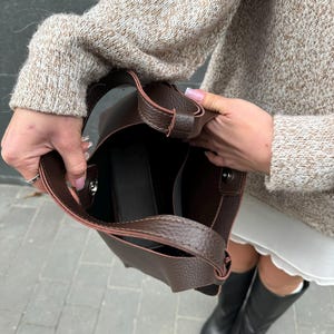 May include: A dark brown leather tote bag with a structured design. The bag has a shoulder strap and a spacious interior. The bag is held open, revealing the inside. The bag is a stylish accessory.