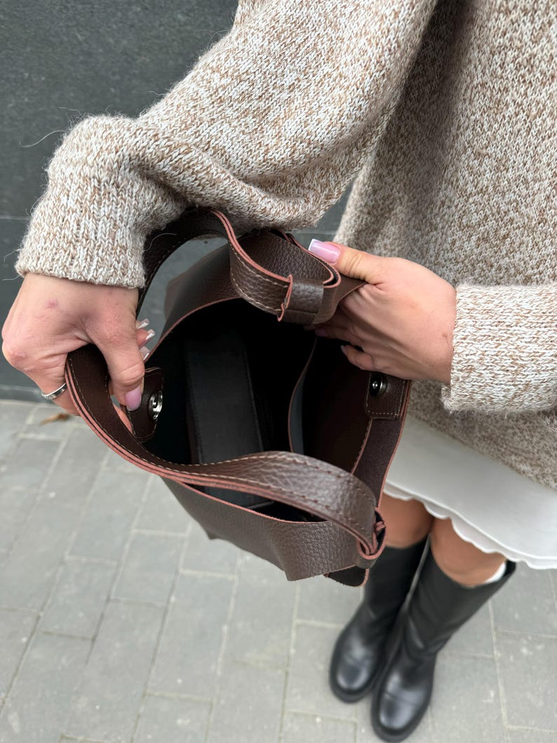 May include: A dark brown leather tote bag with a structured design. The bag has a shoulder strap and a spacious interior. The bag is held open, revealing the inside. The bag is a stylish accessory.