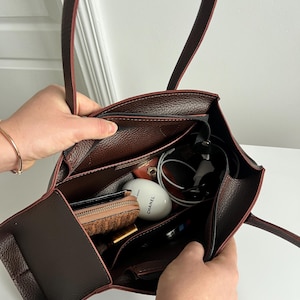 May include: A brown leather tote bag with a structured design, featuring a zippered main compartment and multiple interior pockets. The bag is filled with various items, including a wallet, cosmetics, and a pair of headphones.