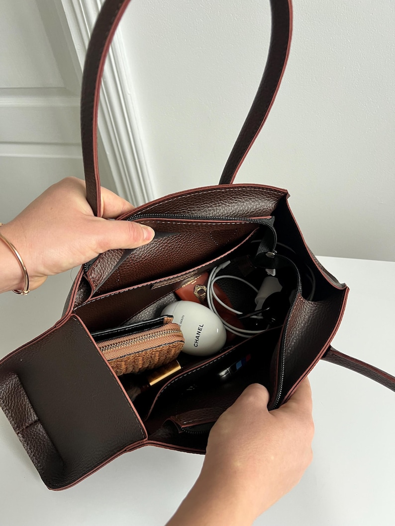 May include: A brown leather tote bag with a structured design, featuring a zippered main compartment and multiple interior pockets. The bag is filled with various items, including a wallet, cosmetics, and a pair of headphones.